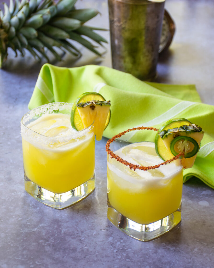 Two pineapple margaritas in rocks glasses with a cocktail shaker and a pineapple top behind them.