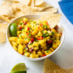 A bowl of pineapple salsa with tortilla chips and lime wedges around.