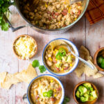 Two small bowls of white chicken chili next to a large stockpot of chili, some small bowls of jalapeño slices and grated cheese.