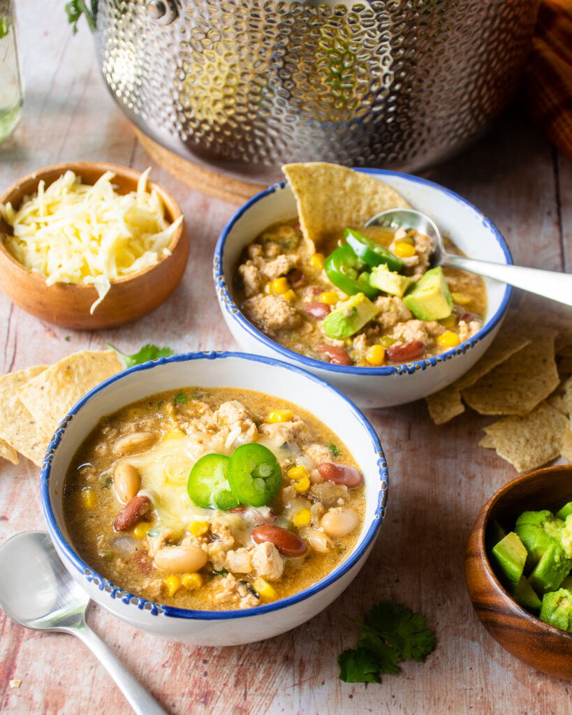 Two blue rimmed white bowls with white chicken chili, grated cheese and Jalapeño pepper on top, all in front of a hammered stainless steel stock pot.