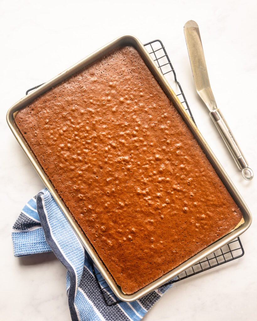 Looking down on a chocolate sheet cake on a cooling rack with an offset spatula and a blue striped towel on the side.