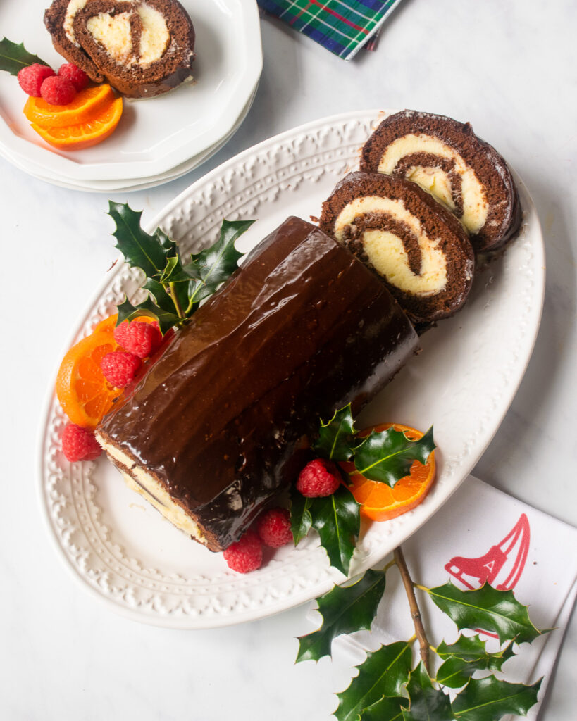 a chocolate coated Chocolate Swiss Roll on a white plate with mandarin orange slices, raspberries and holly decorating it and a couple of slices cut.