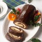 a chocolate coated Chocolate Swiss Roll on a white plate with mandarin orange slices, raspberries and holly decorating it and a couple of slices cut.