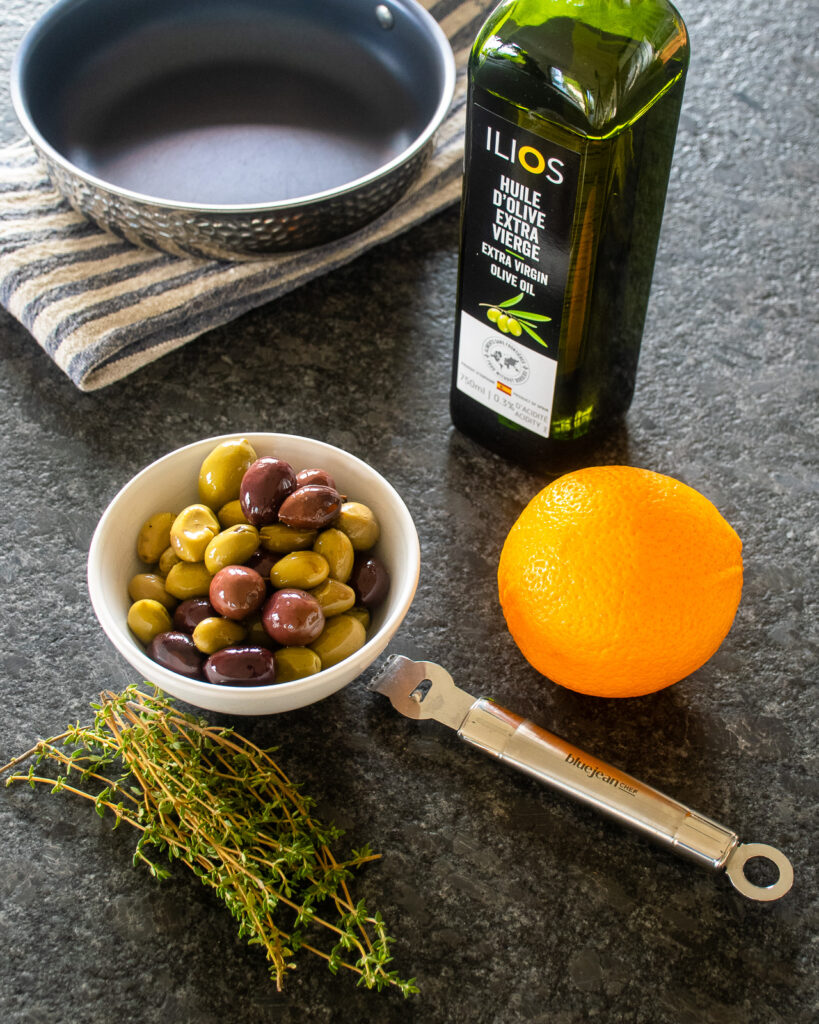 A bowl of olives, an orange, some fresh thyme, a bottle of olive oil and a channel knife on a counter.