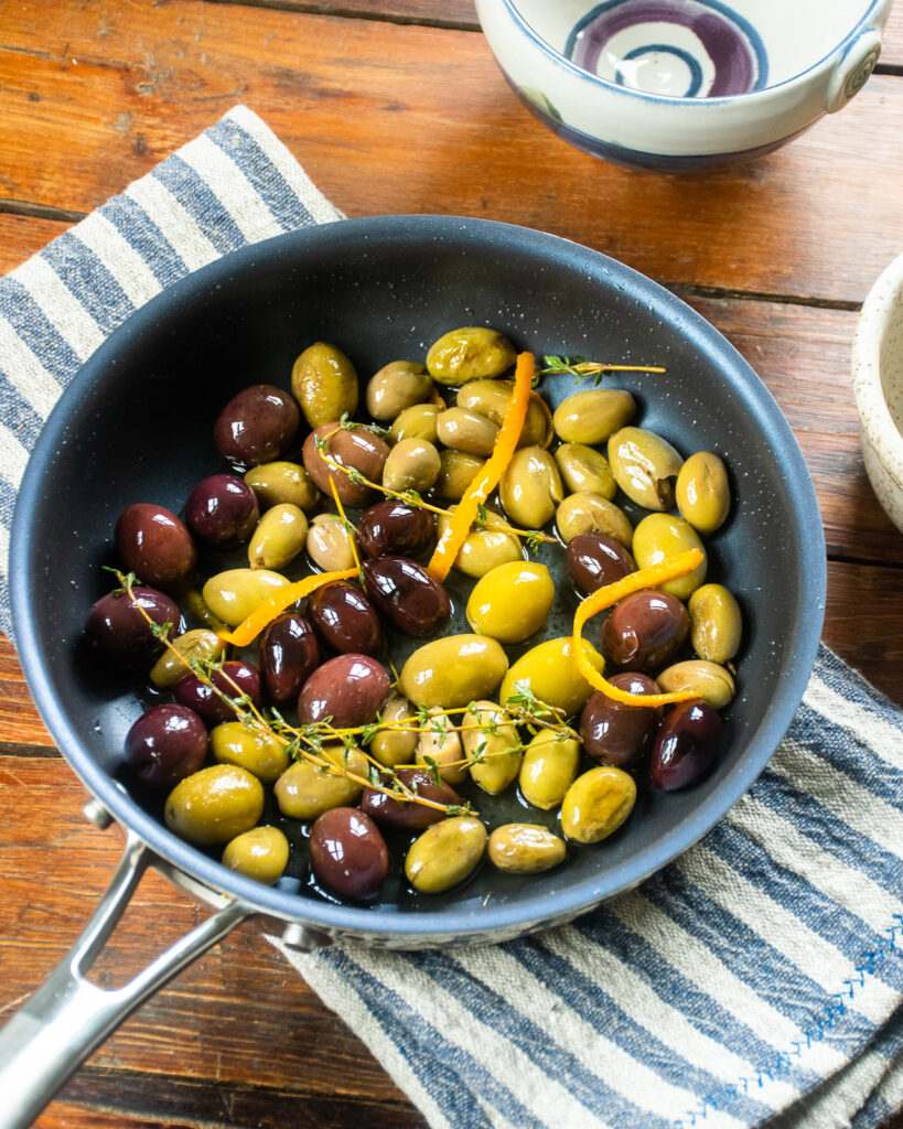 A skillet with olives, orange peel, fresh thyme on a wooden counter top with a blue and grey striped kitchen towel.