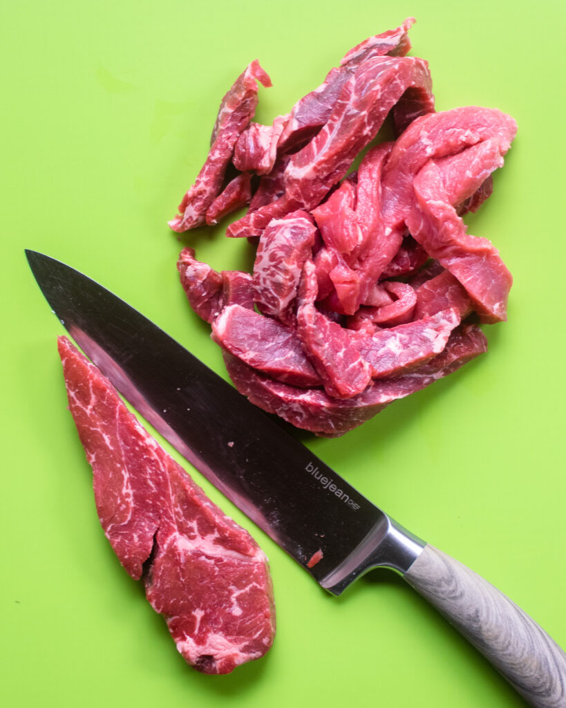 Sirloin steak being cut into strips with a chef's knife on a bright green cutting board.