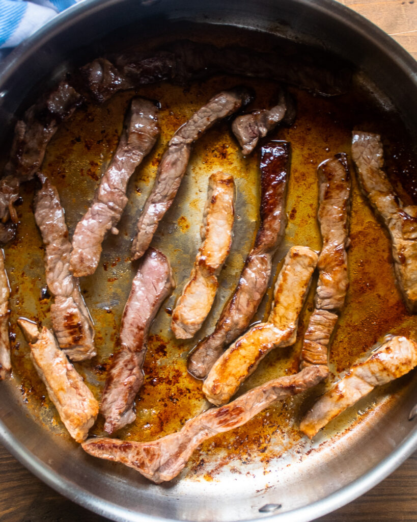 Strips of beef browning in a stainless steel sauté pan.