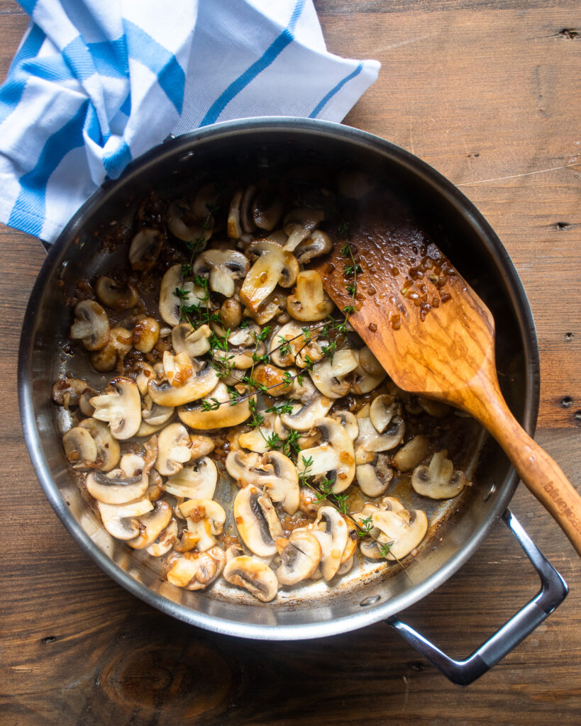 Mushrooms and thyme in a sauté pan with a wooden spoon sticking out of it and a blue and white striped towel on one handle.