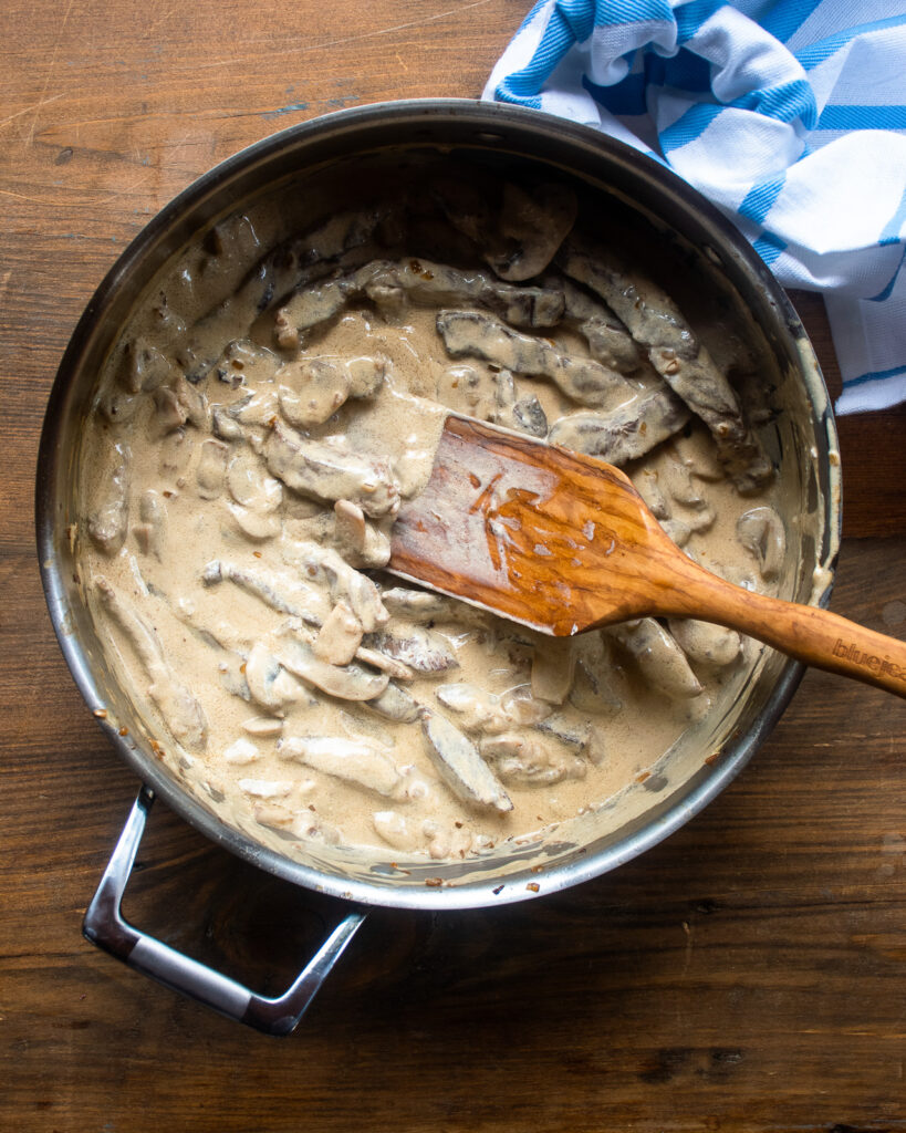 Beef stroganoff in a sauté pan with a wooden spoon sticking out of it and a blue and white kitchen towel over one handle of the pan.