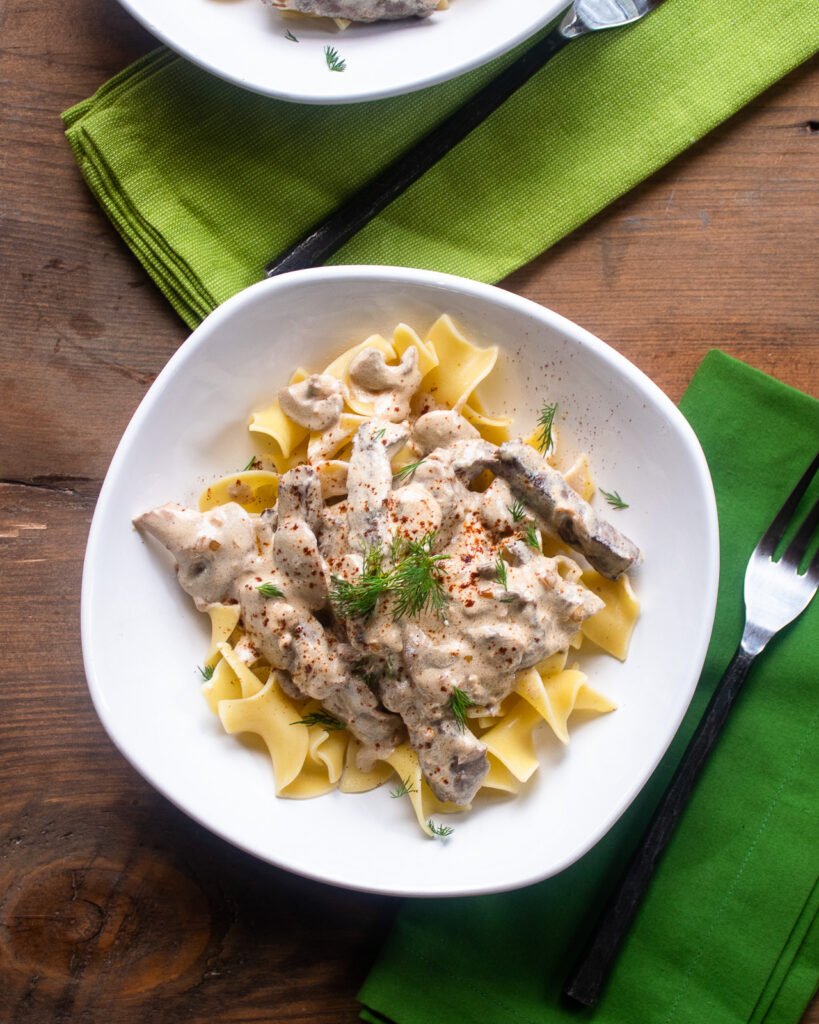 Looking down on a white bowl with beef stroganoff inside and a green napkin with fork.