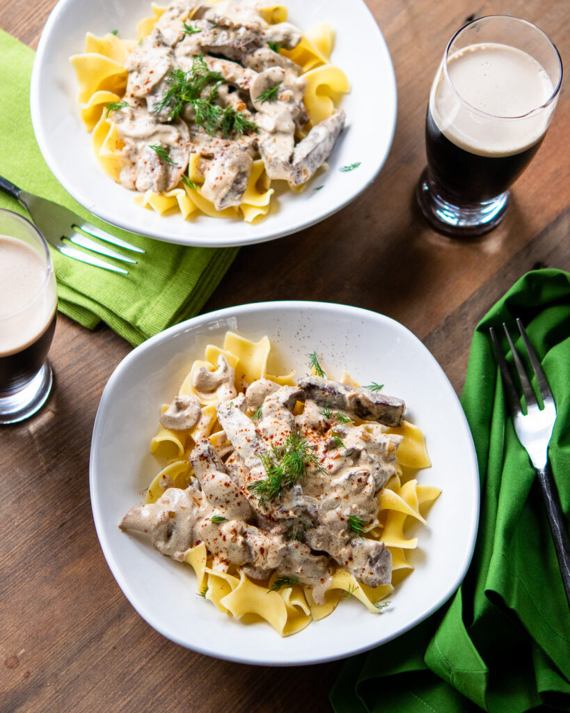 Two bowls of beef stroganoff in white bowls over egg noodles with a glasses of Guinness.