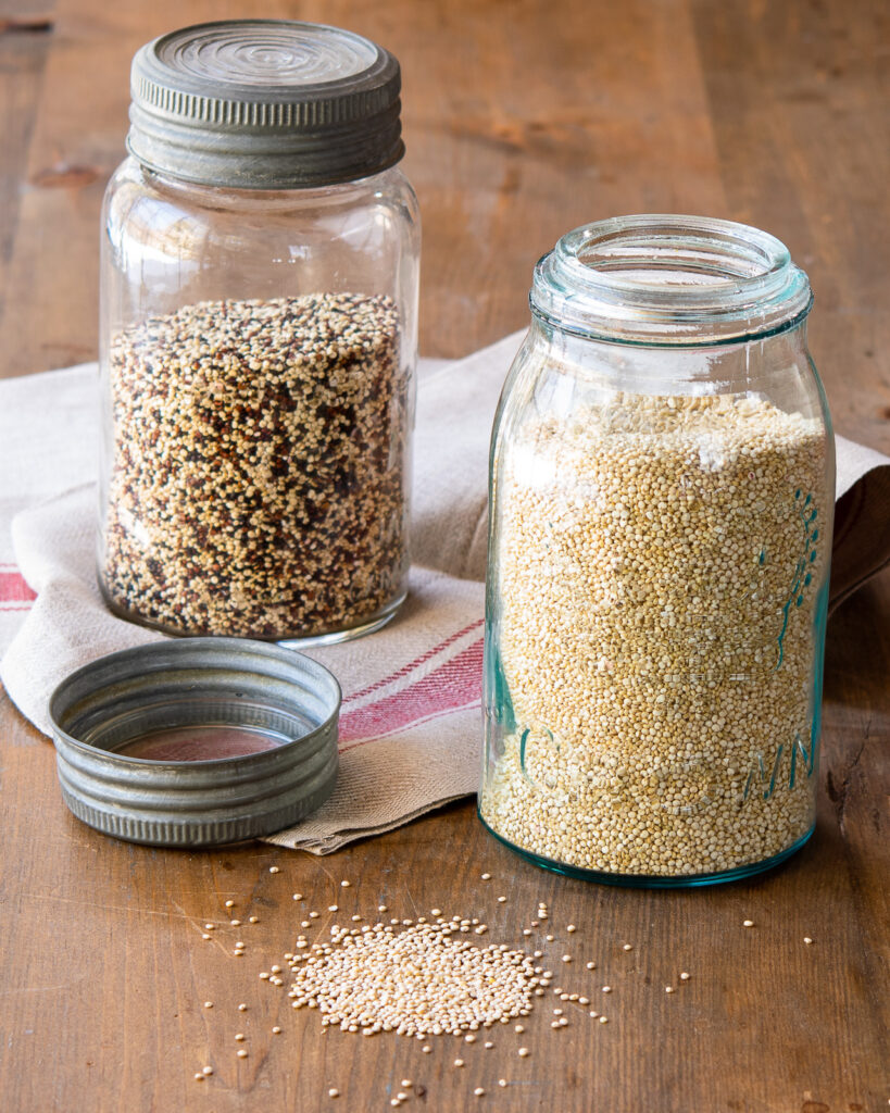 Two jars of quinoa - one white and one multi-colored - on a wooden table with a small pile of white quinoa in the front.
