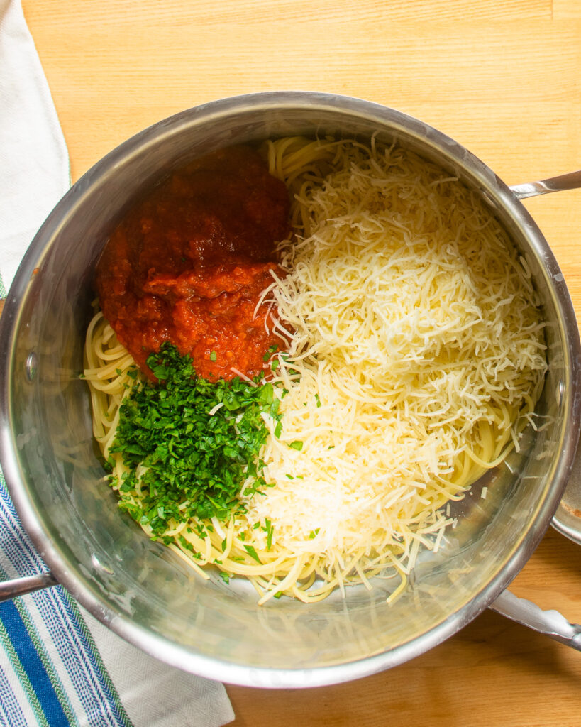 Looking down into a stockpot with spaghetti, marinara sauce, cheeses and parsley.