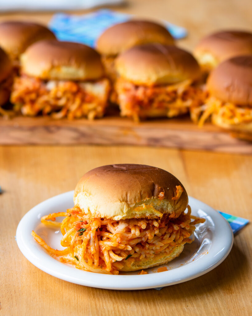 A spaghetti slider on a small plate in front of a wooden board with more sliders on it.